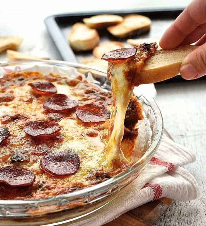 Toasted bread scooping up ultra cheesy, gooey Pizza Dip in a glass dish with a great cheese pull shot.